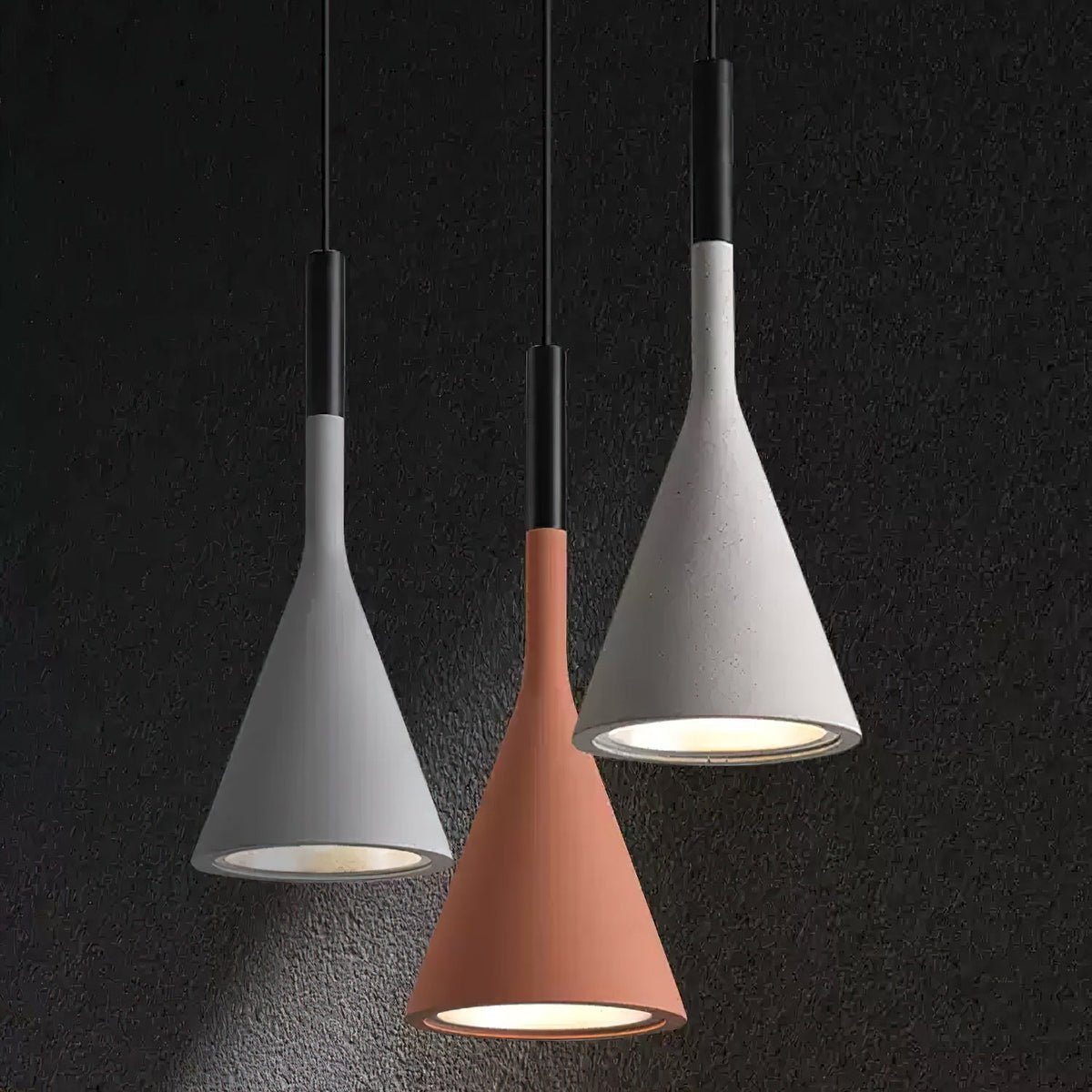 Three Clérachic concrete pendants in two greys and a terracotta orange against a dark textured wall, each lit with a silver-lined opening.
