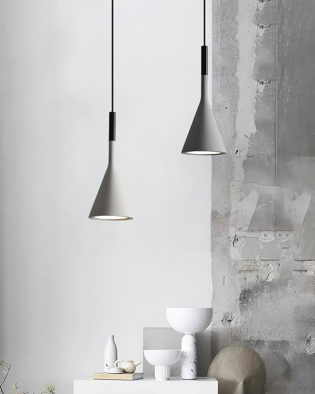 Two grey concrete Clérachic pendants against a weathered grey wall, above a white shelf with bowls and a stone sculpture.