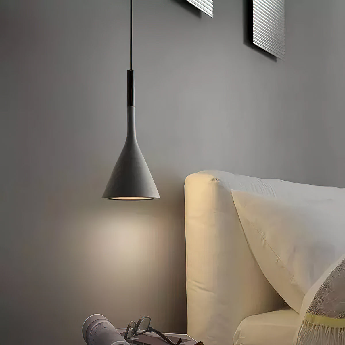 Dark grey concrete Clérachic pendant against a grey wall, hung beside a cream upholstered headboard with a folded throw.