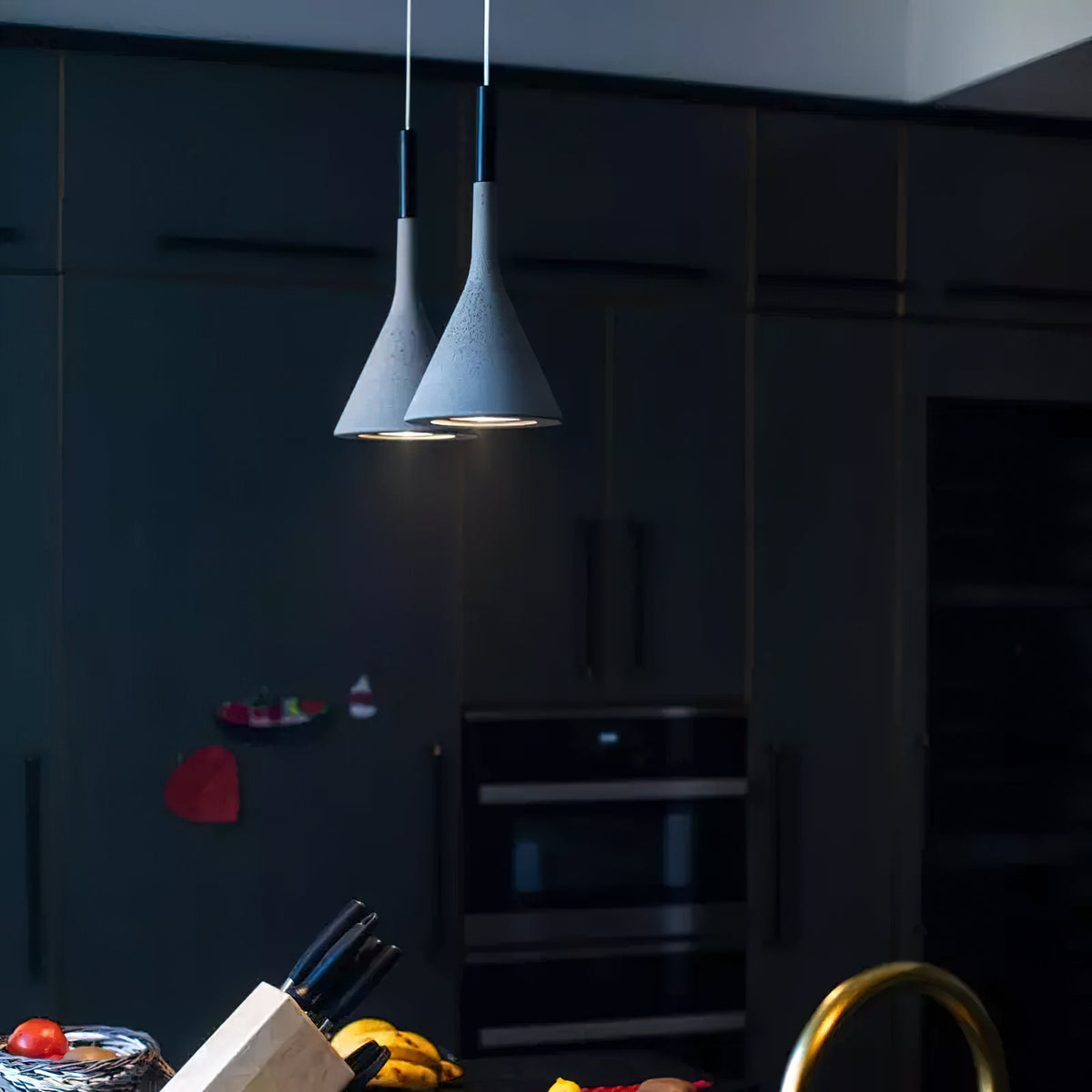 Two blue-grey concrete Clérachic pendants lit in a dark kitchen of black cabinets, with a brass tap in the foreground.