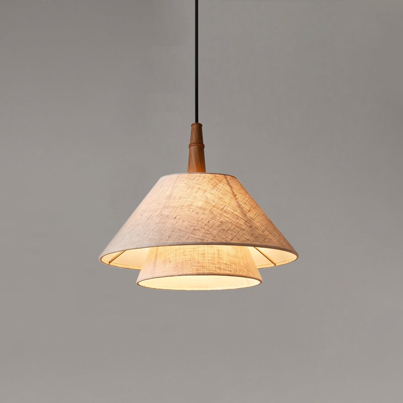 A lit Clérachic pendant with two linen tiers and a wooden neck, hung on a black cord against a mid-grey wall.