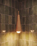 Close view of a Clérachic oak-grain wooden cone pendant with a brass collar and a lit globe bulb against green tiles.