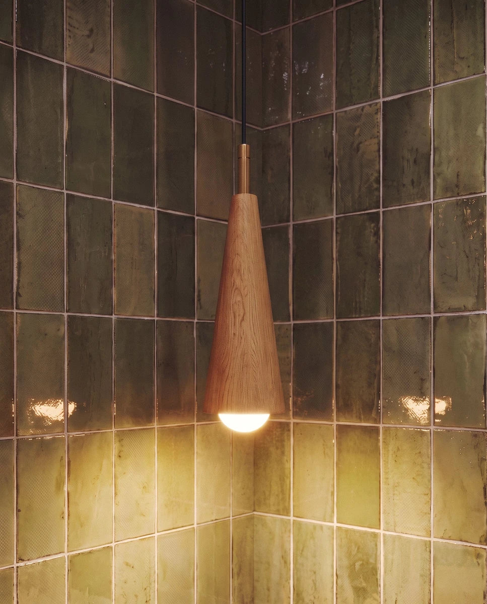 Close view of a Clérachic oak-grain wooden cone pendant with a brass collar and a lit globe bulb against green tiles.