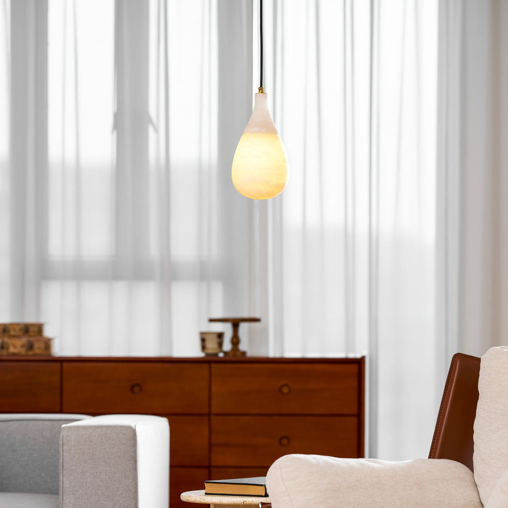 Lit warm, a small Clérachic alabaster drop hangs on a black cord in front of sheer curtains and a walnut sideboard.