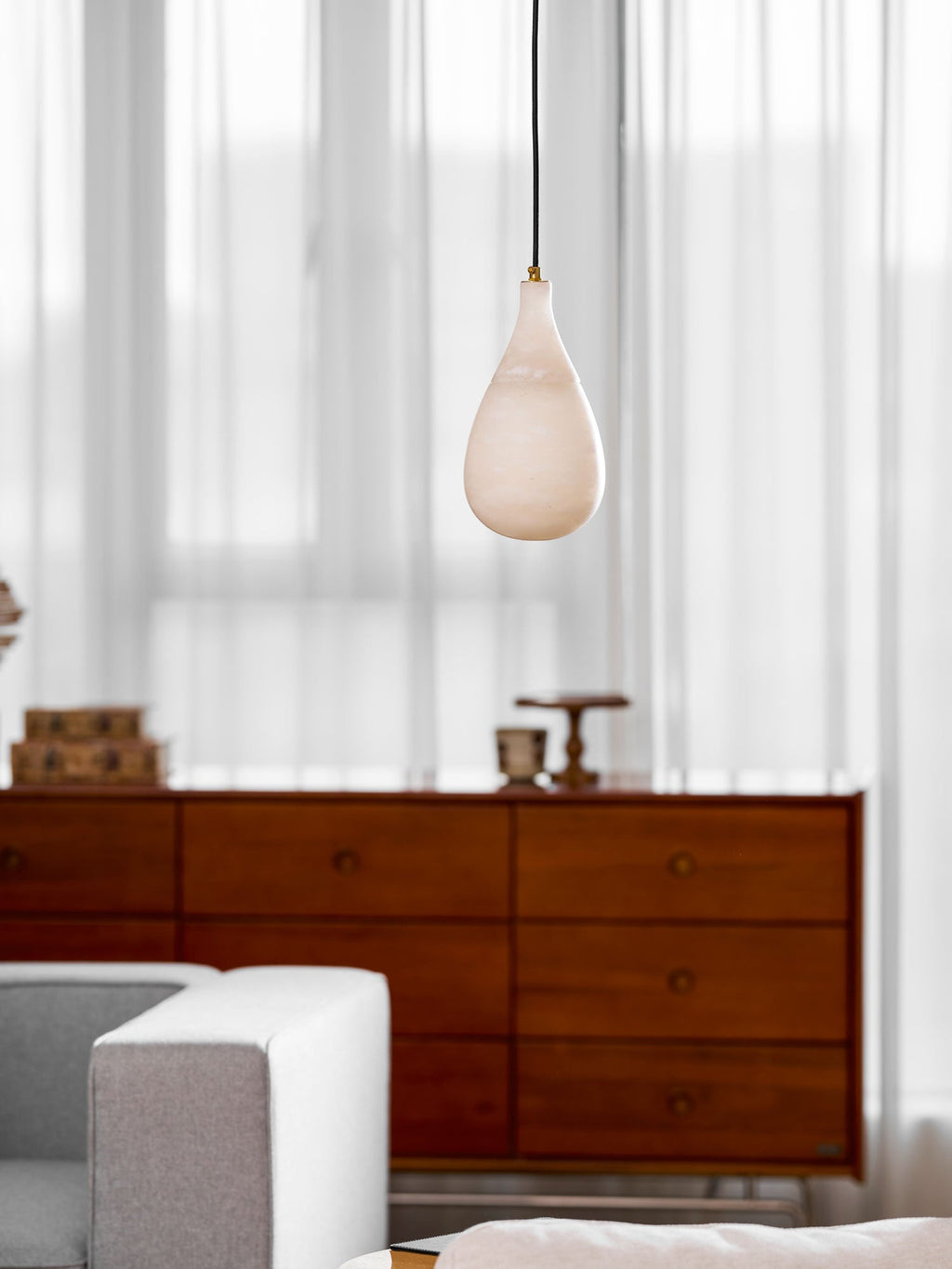 The unlit alabaster shade of this Clérachic pendant hangs in front of sheer curtains, with a walnut sideboard below.