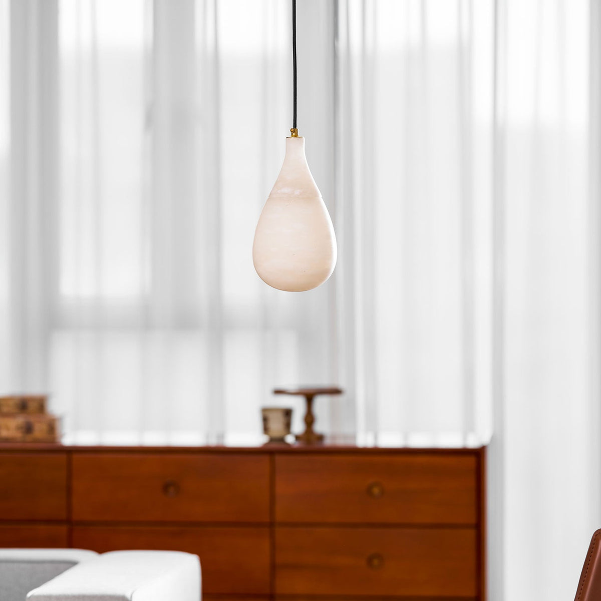 Small unlit Clérachic alabaster pendant on a black cord, with a walnut chest of drawers and curtains behind.