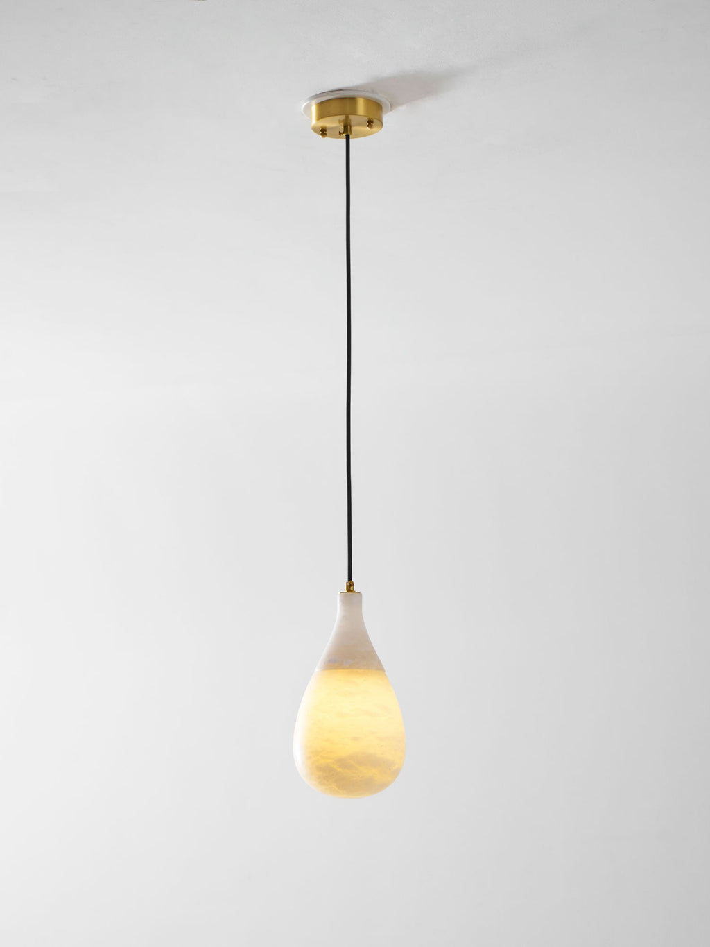 A lit Clérachic alabaster drop hangs on a long black cord from a round brass ceiling plate, against a pale wall.