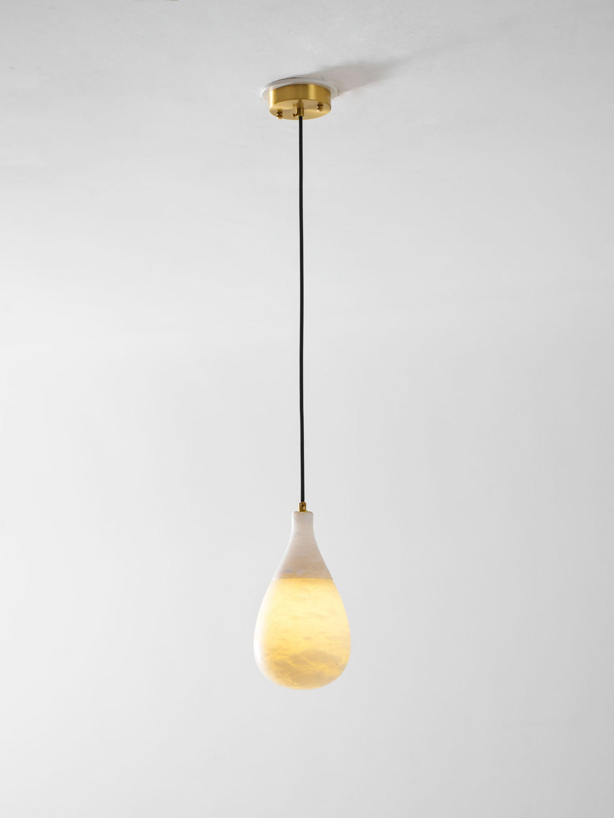 A lit Clérachic alabaster drop hangs on a long black cord from a round brass ceiling plate, against a pale wall.