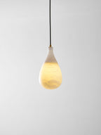 A lit Clérachic alabaster drop on a black cord, glowing warm in its lower half against a plain grey wall.