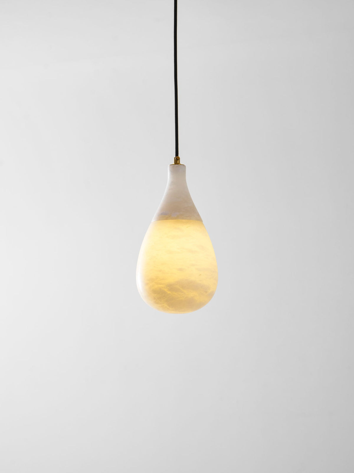 A lit Clérachic alabaster drop on a black cord, glowing warm in its lower half against a plain grey wall.