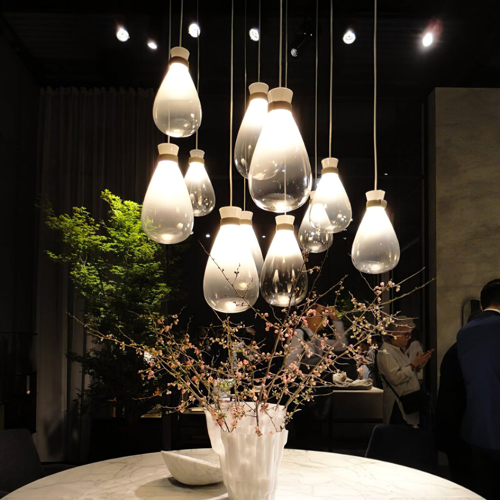 Cluster of Clérachic teardrop glass pendants, some frosted and some clear, glowing over a round table with a vase of blossom in a dark room.