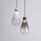 A white and a smoke-grey Clérachic teardrop pendant side by side against a pale grey wall, each with a leather strap.