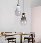 A clear and a smoke-grey Clérachic teardrop pendant hanging over a wooden dining table with grey chairs and a pink wall.