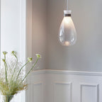 Clérachic teardrop pendant with a frosted top and clear base, hanging in a panelled room beside a vase of white flowers.