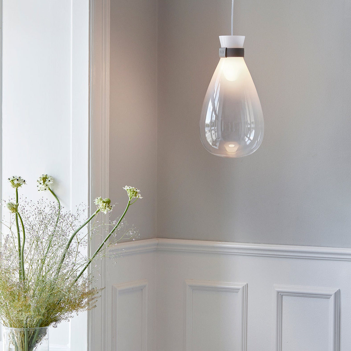 Clérachic teardrop pendant with a frosted top and clear base, hanging in a panelled room beside a vase of white flowers.