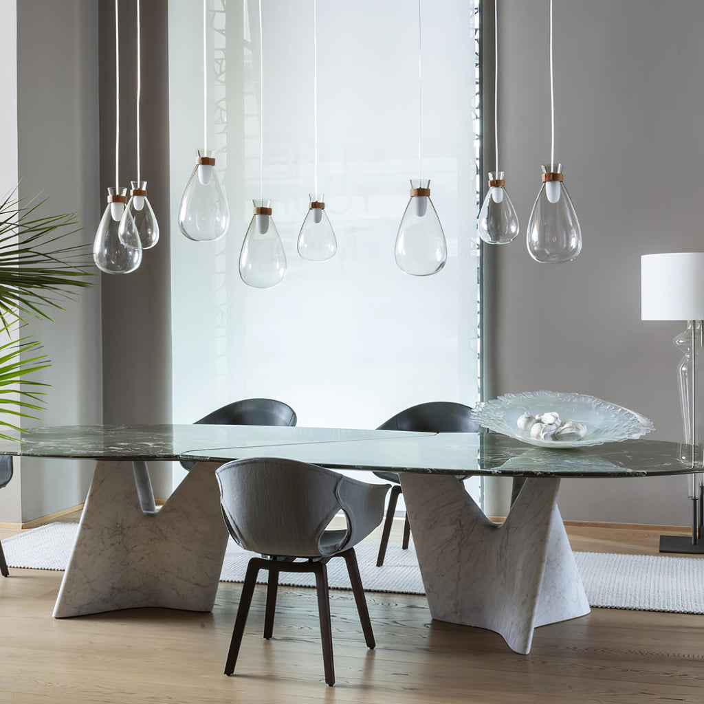 Clear glass Clérachic teardrop pendants at mixed heights over a long glass-topped table with stone legs and grey chairs.