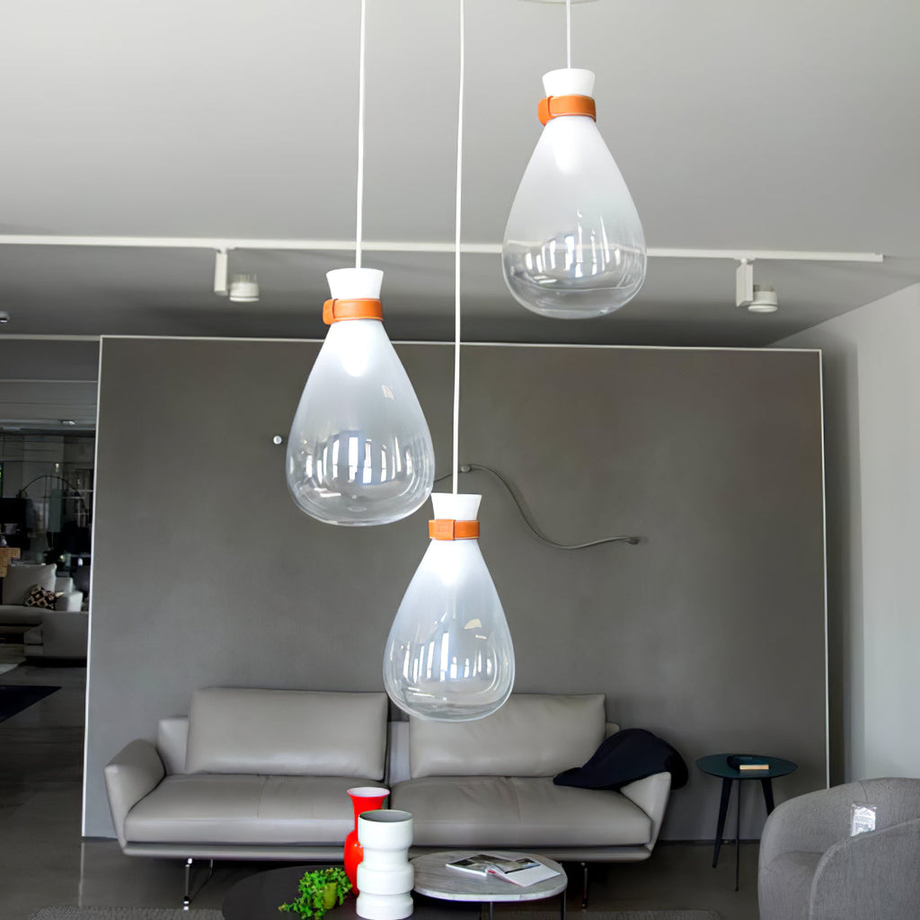 Three clear and frosted Clérachic teardrop pendants with leather straps, over a grey sofa in a showroom with track lights.