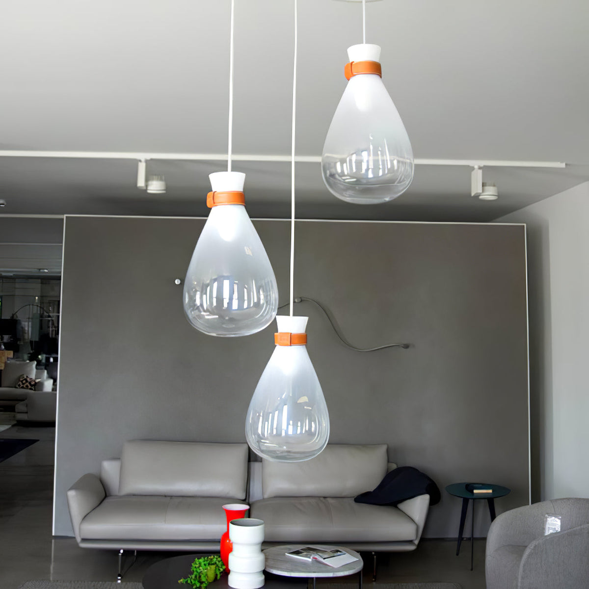 Three clear and frosted Clérachic teardrop pendants with leather straps, over a grey sofa in a showroom with track lights.