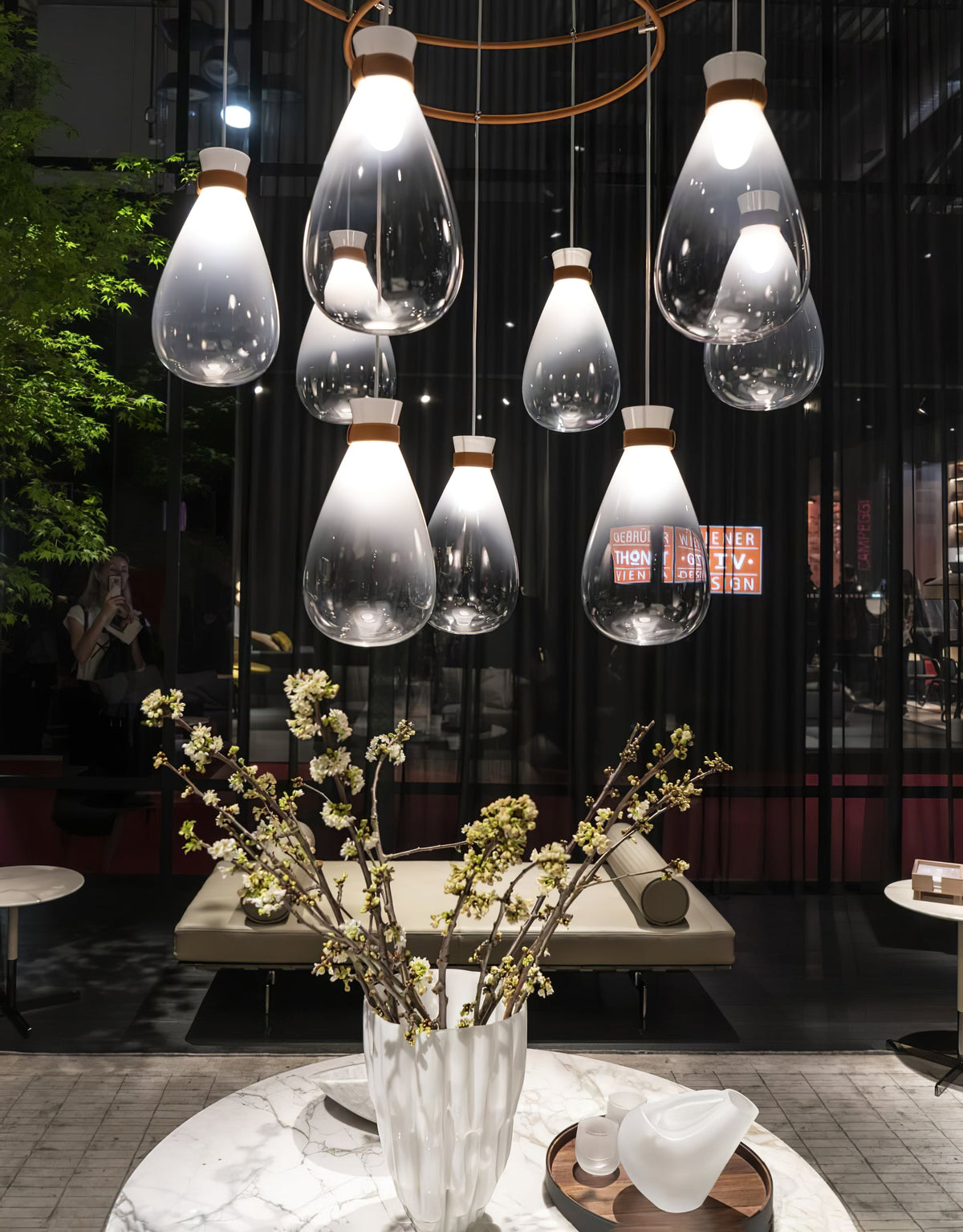 Clérachic teardrop pendants of varied sizes hung in a cluster over a table with a vase of yellow blossom, in a dark showroom.