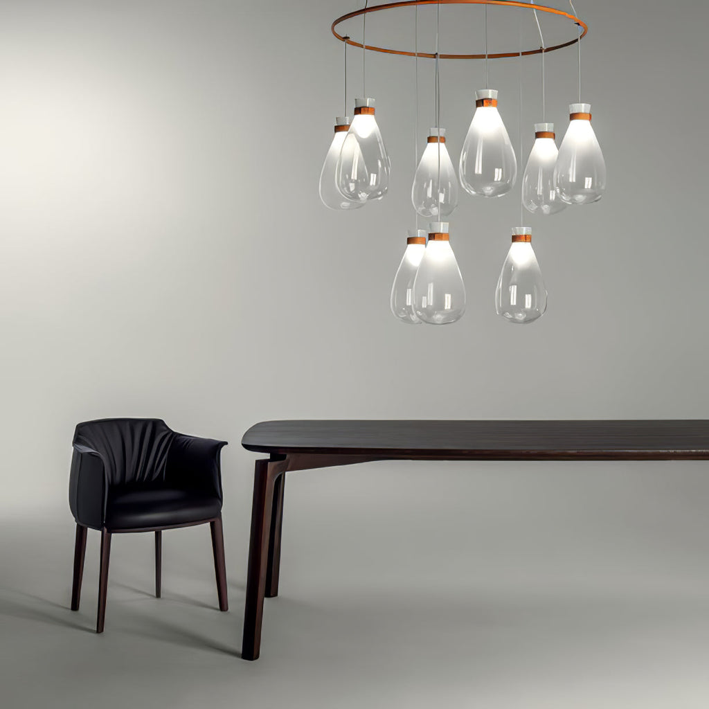 Clear glass Clérachic teardrop pendants hung from a copper ring over a dark table and a black armchair on a pale grey backdrop.