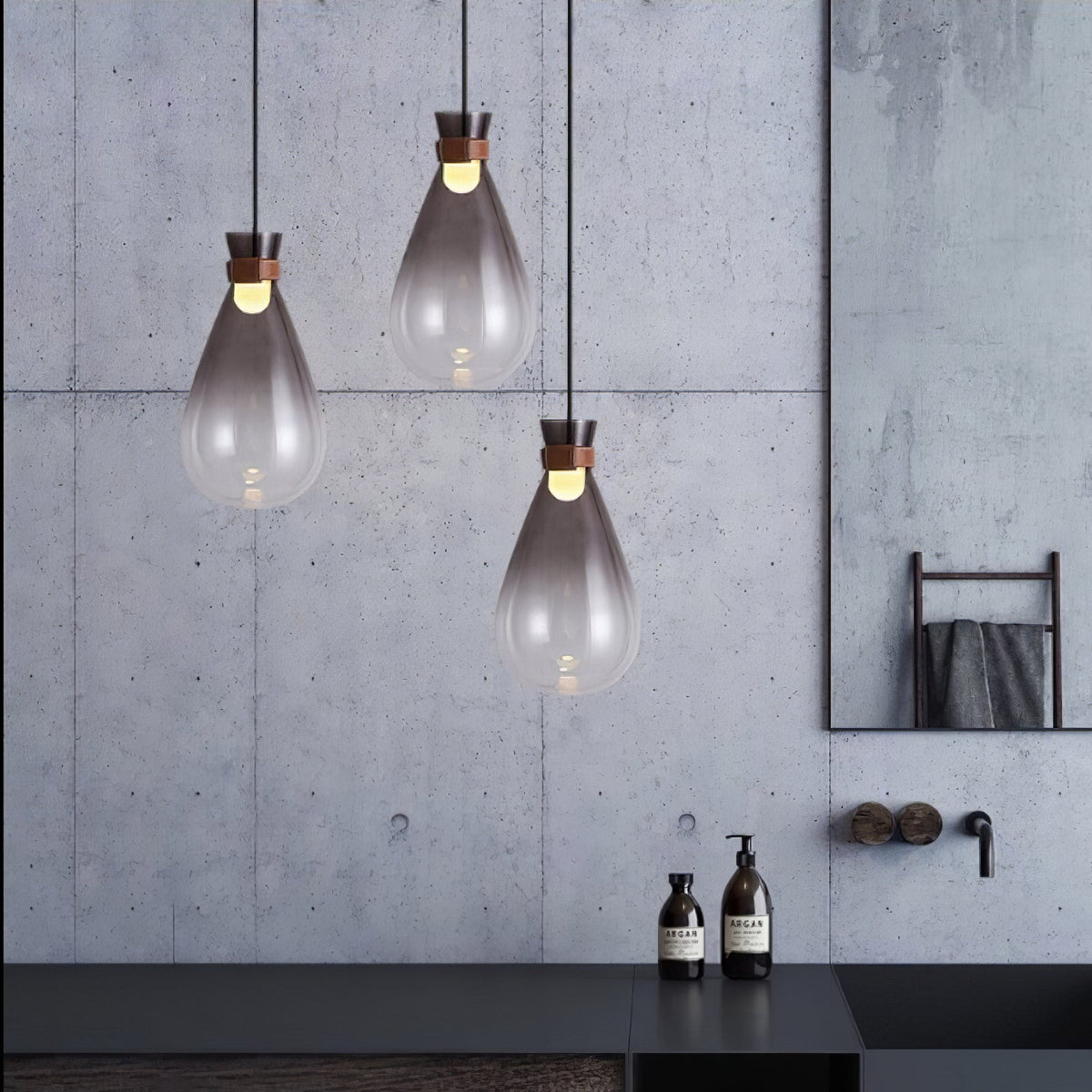 Three smoke-grey Clérachic teardrop pendants against a concrete wall, with a dark counter and soap bottles below.