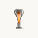 This Clérachic table lamp on a plain white backdrop: a smoke-grey glass teardrop shade around a glowing orange core, on a round chrome base.