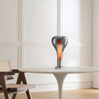 On a white pedestal table beside a rattan-seat wooden chair, a smoke-grey teardrop lamp glows with a warm core, from Clérachic.