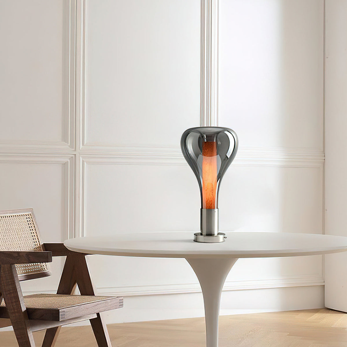 On a white pedestal table beside a rattan-seat wooden chair, a smoke-grey teardrop lamp glows with a warm core, from Clérachic.