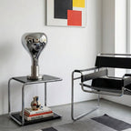 A mirror-finish teardrop shade on a chrome base sits on a tubular steel side table beside a leather-and-steel armchair, from Clérachic.