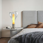 A clear teardrop glass lamp with a glowing amber column sits on a round stone side table by a grey upholstered bed, from Clérachic.