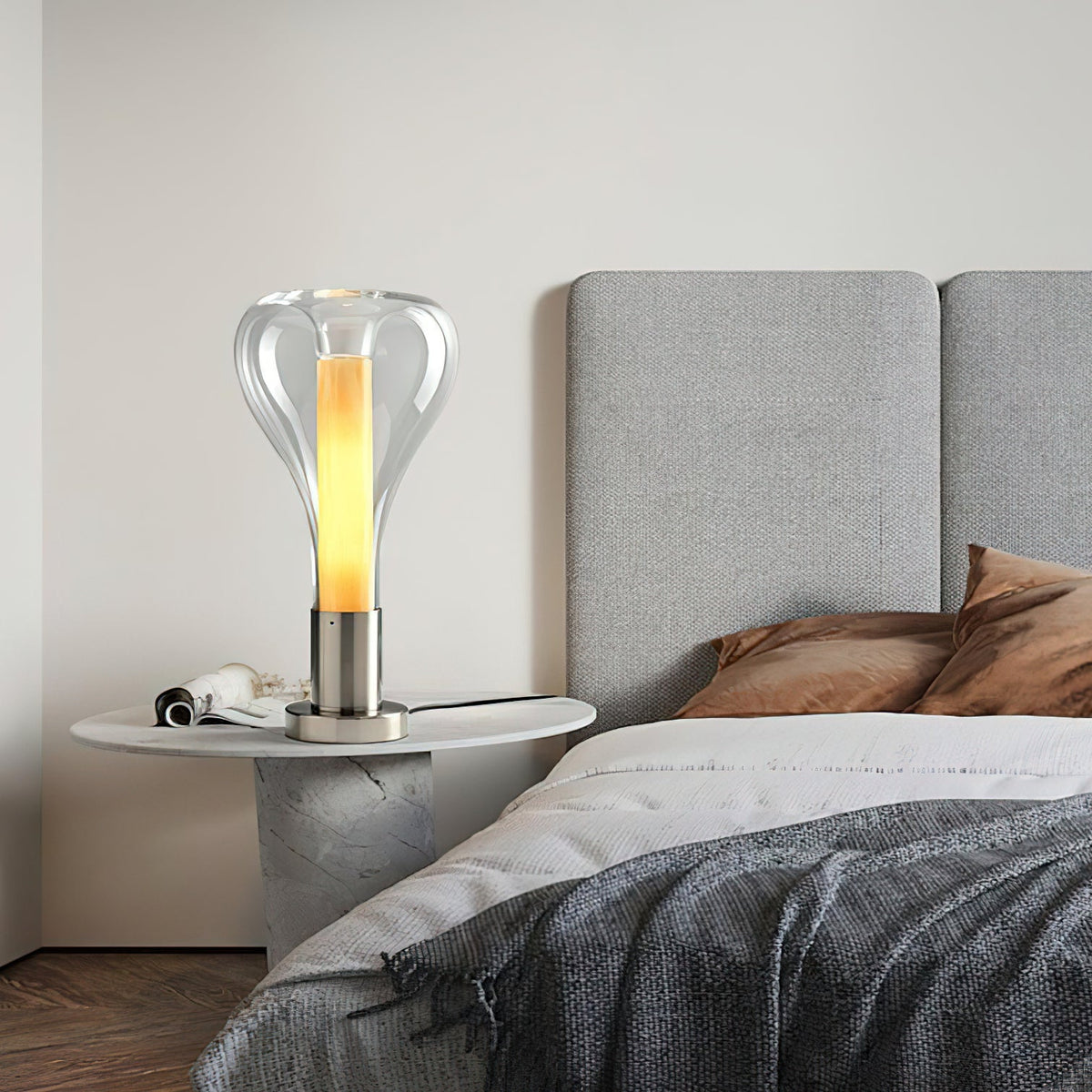 A clear teardrop glass lamp with a glowing amber column sits on a round stone side table by a grey upholstered bed, from Clérachic.