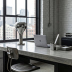 On a concrete desk by a tall industrial window, a mirror-chrome teardrop lamp stands beside a laptop and pen pot, from Clérachic.
