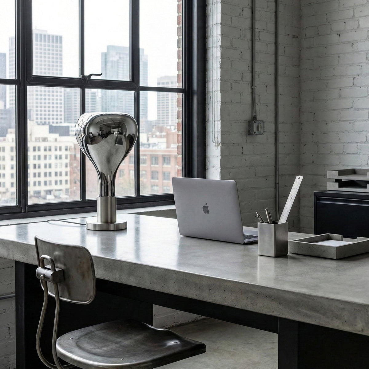 On a concrete desk by a tall industrial window, a mirror-chrome teardrop lamp stands beside a laptop and pen pot, from Clérachic.