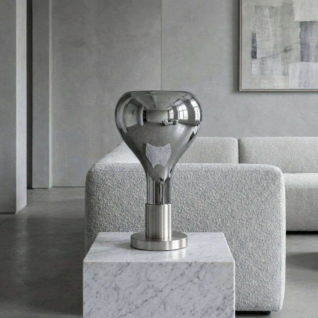 A mirrored teardrop lamp with a chrome stem stands on a white marble block in front of a pale boucle sofa, from Clérachic.