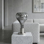 A mirrored teardrop lamp with a chrome stem stands on a white marble block in front of a pale boucle sofa, from Clérachic.