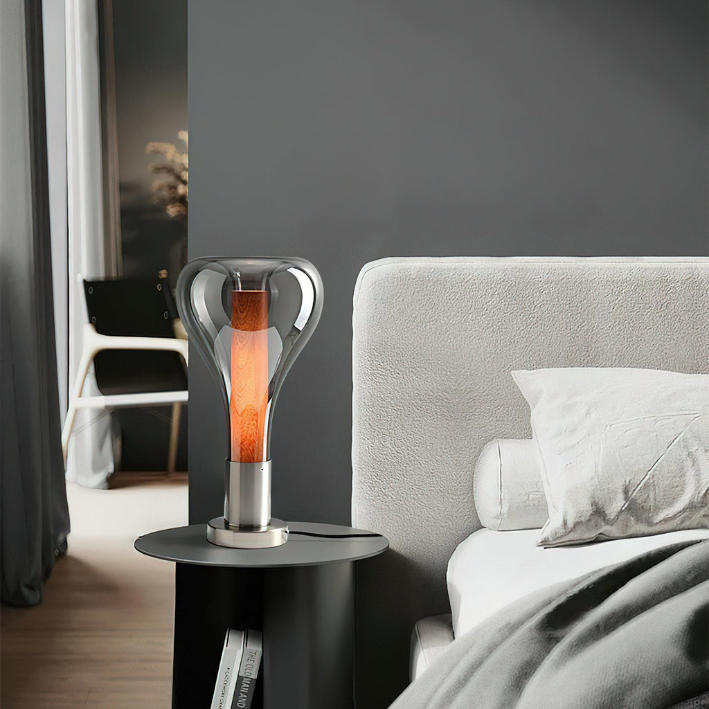 Beside a bed with white bedding, a smoke-grey teardrop lamp glows with an amber core on a round black table, from Clérachic.