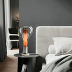 Beside a bed with white bedding, a smoke-grey teardrop lamp glows with an amber core on a round black table, from Clérachic.