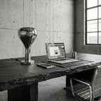 A mirror-chrome teardrop lamp stands on a dark slate desk beside an open laptop, against a raw concrete wall, from Clérachic.