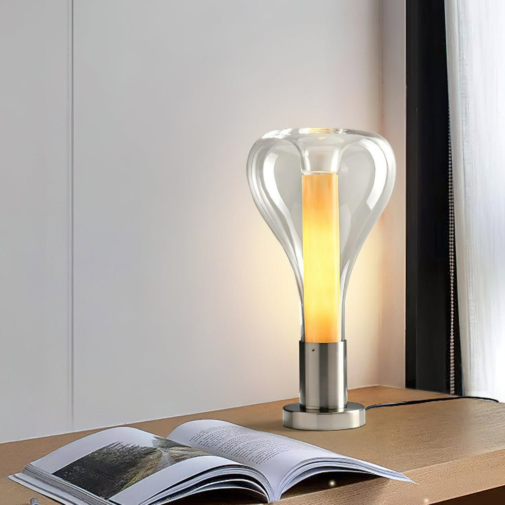 A clear glass teardrop lamp with a glowing amber core stands on a wooden shelf beside an open book and a curtain, from Clérachic.