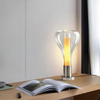 Clear glass teardrop lamp, lit amber, beside an open book on a light wood shelf, with a curtain to the right, from Clérachic.