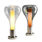 Cut out on white, a clear lit teardrop lamp stands beside a smoke-grey one with an amber core, both on round chrome bases, from Clérachic.