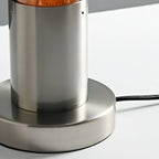 Close view of the brushed metal stem and round base on this Clérachic lamp, with its black cord leaving the base on a white surface.