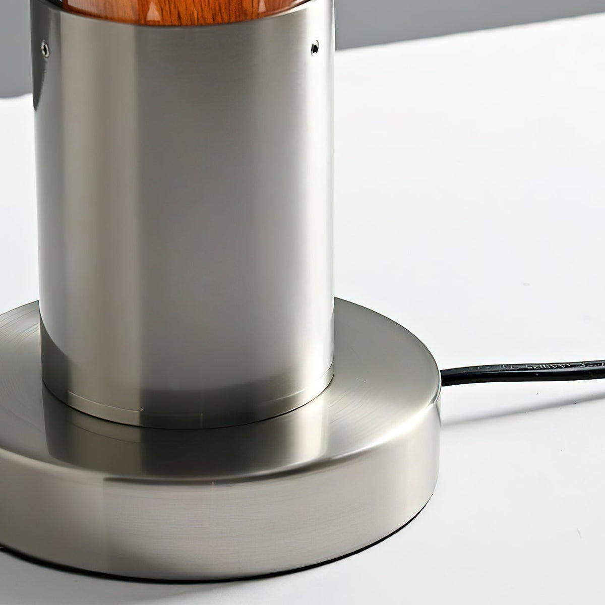 Close view of the brushed metal stem and round base on this Clérachic lamp, with its black cord leaving the base on a white surface.