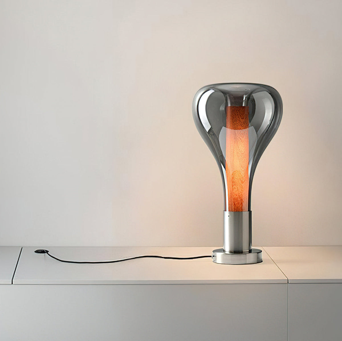 A smoke-grey teardrop lamp with a glowing wood-toned core stands on a white sideboard with its black cord trailing, from Clérachic.