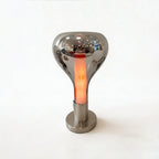 A mirror-chrome teardrop shade with a warm pink-orange glow inside, on a brushed round base against a light grey wall, from Clérachic.