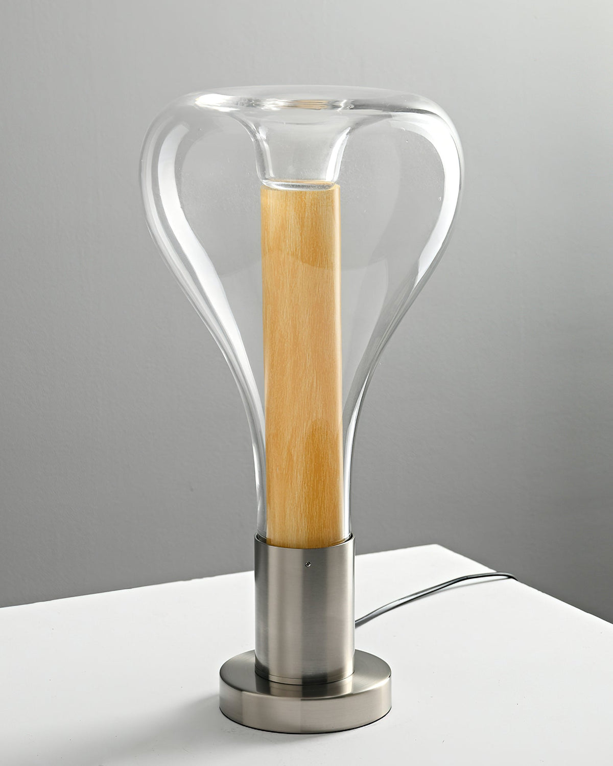 A clear glass teardrop lamp with a pale wooden core on a brushed metal base, standing on a white shelf against a grey wall, from Clérachic.