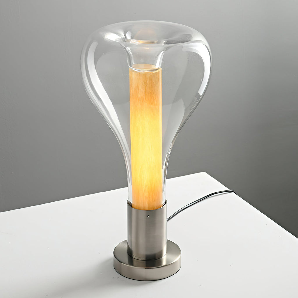 Lit warm amber, a clear teardrop lamp stands on a white shelf with its cord trailing behind, against a grey wall, from Clérachic.