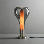 A smoke-grey teardrop lamp with a deep orange wooden core stands on a brushed metal base on a grey surface, from Clérachic.