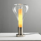 A clear teardrop lamp with a lit amber column on a chrome base, standing on a white shelf against grey, from Clérachic.