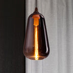 Clérachic teardrop pendant in smoke gray glass, filament lit, against a linen curtain and dark wood panel.